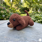 A small crocheted dachshund toy lying on a gray stone table outdoors, with a blurred garden background of green foliage and flowers.