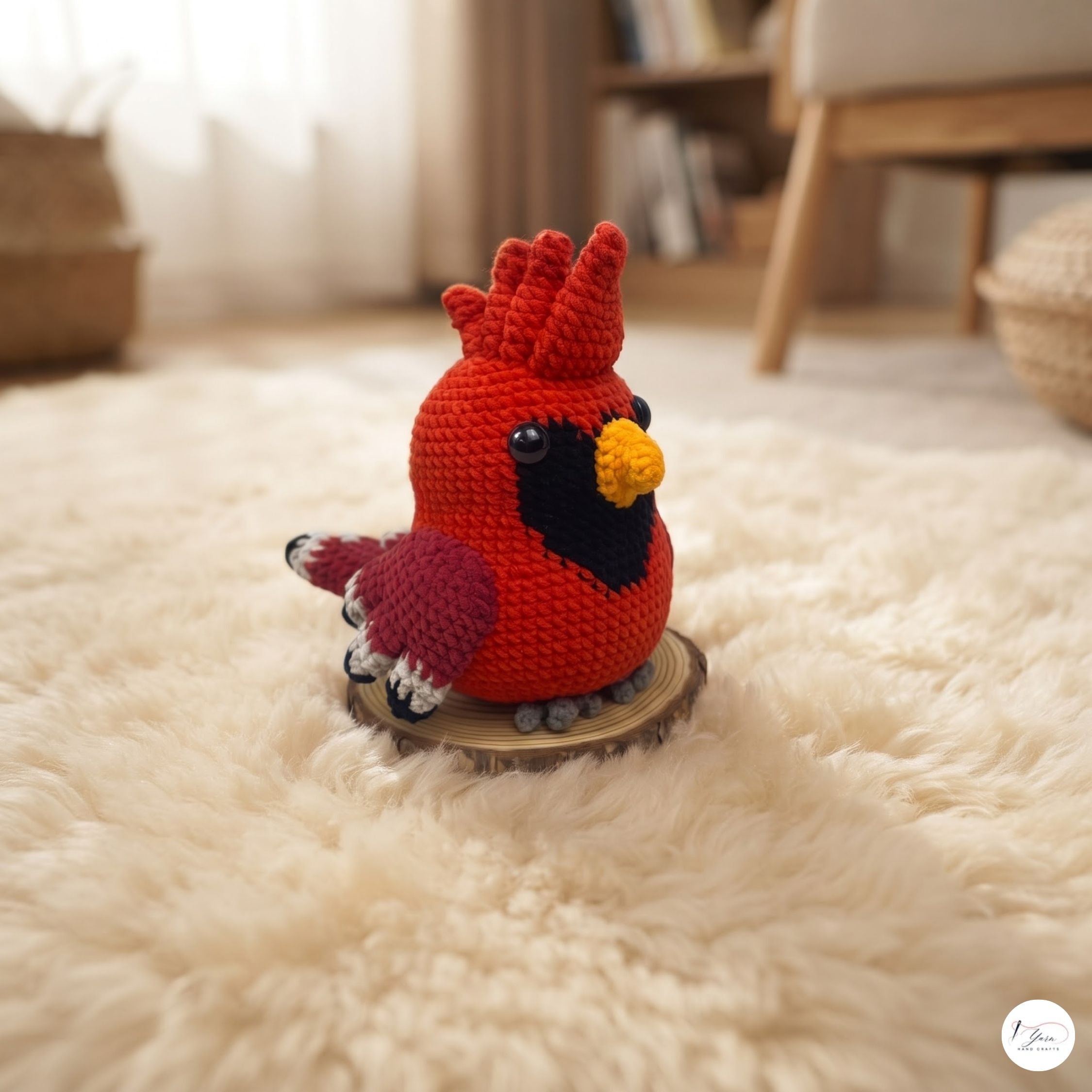 A handmade red cardinal amigurumi toy with a yellow beak, black chest, and pinkish-purple wings, sitting on a small wooden slice coaster on a fluffy white rug in a cozy room.