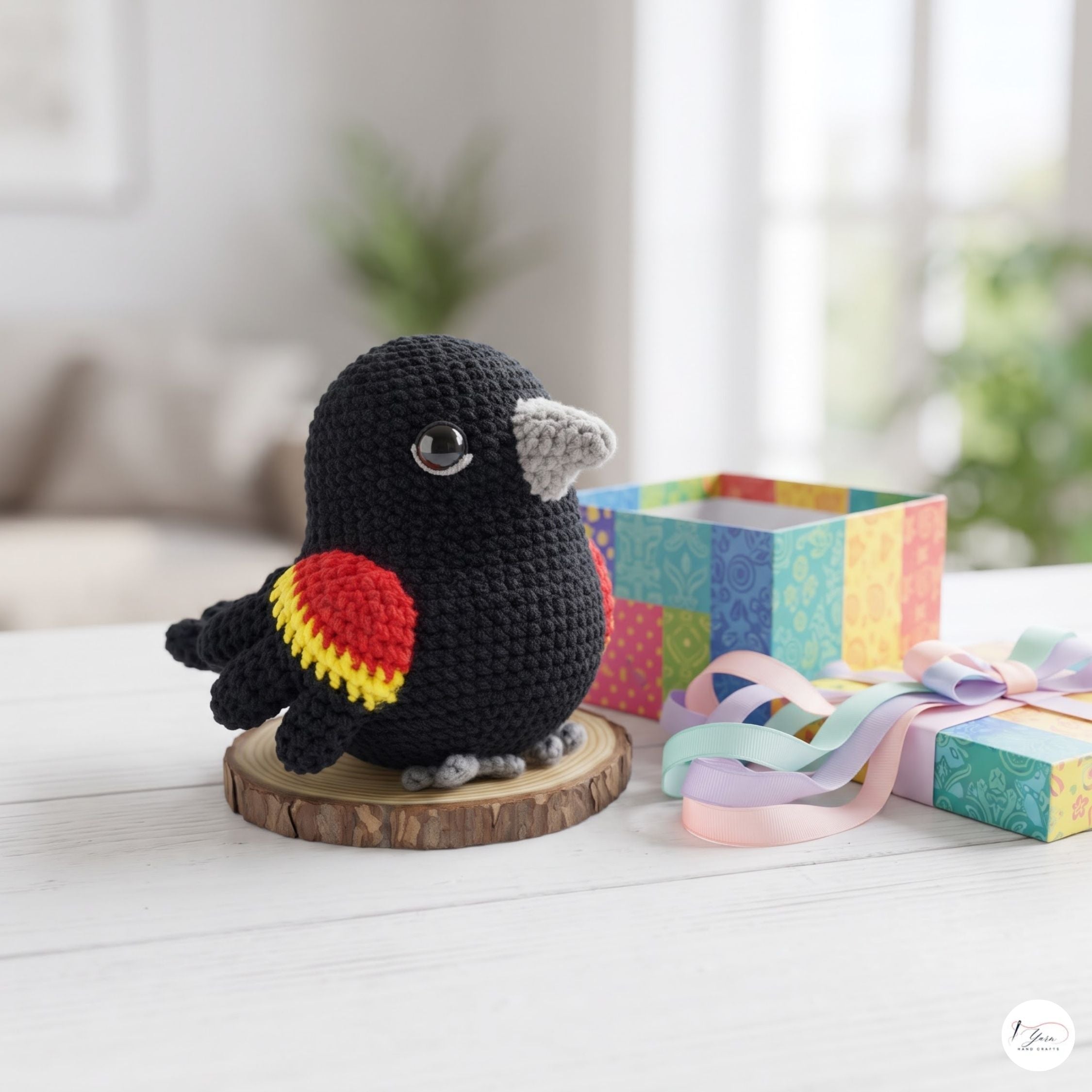 A crocheted black bird figurine with red and yellow wing accents, gray beak and feet, displayed on a wooden slice coaster next to a colorful gift box and pastel ribbon in a bright, softly blurred indoor setting.