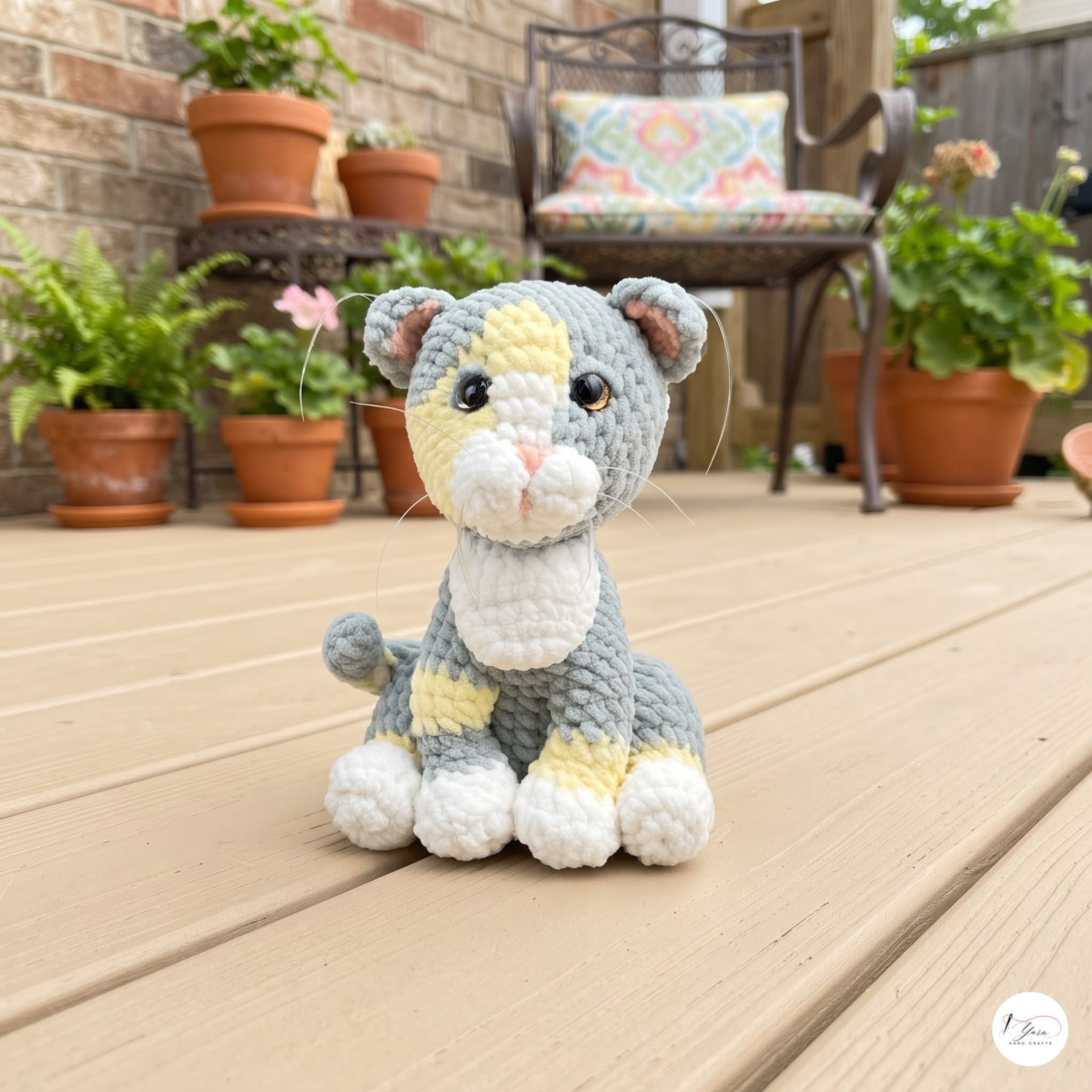 A crocheted cat toy sits on a wooden deck in front of a patio chair and potted plants. The toy is gray with yellow and white patches, black eyes, and a pink nose.