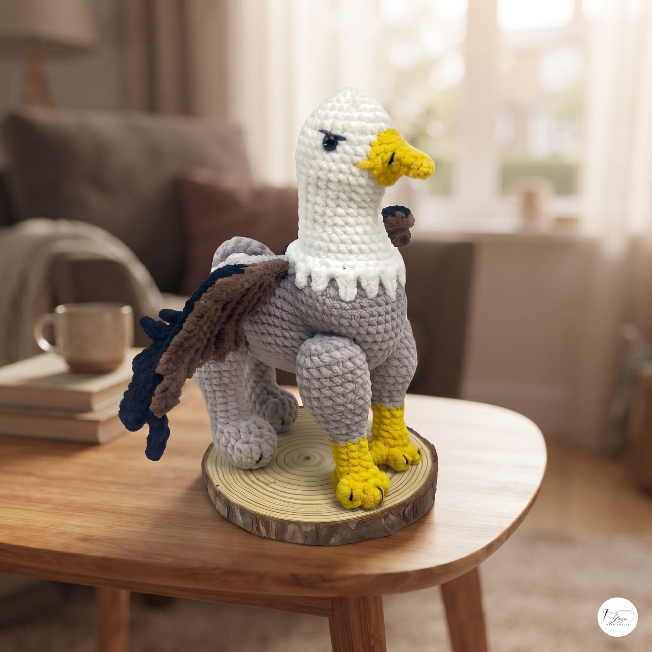 A crocheted eagle figurine standing on a round wood slice base, placed on a wooden table in a softly blurred living room setting.