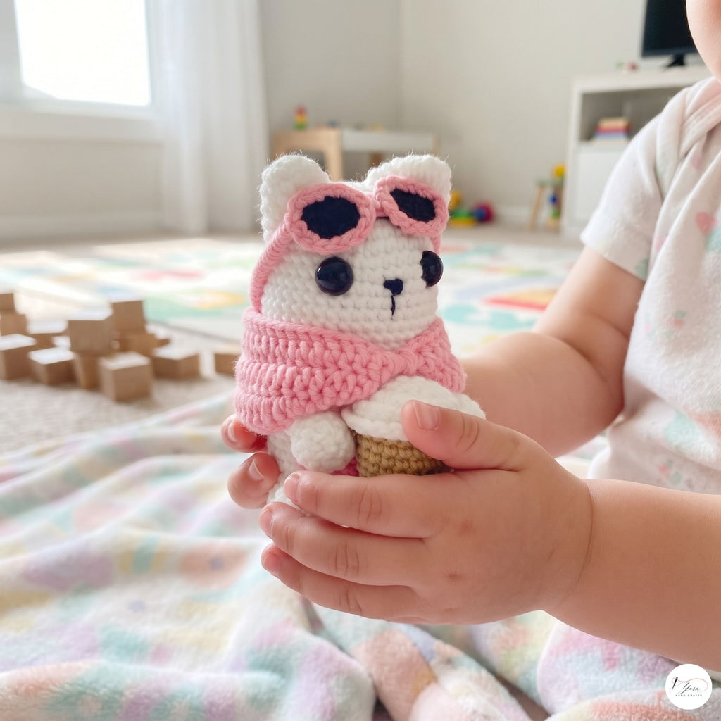A side-profile view of a cute handmade white crochet cat plush sitting on green grass in an outdoor garden, wearing a pink sweater, pink-framed sunglasses over its eyes, holding a crocheted coffee cup, and showing its white tail.