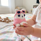 A side-profile view of a cute handmade white crochet cat plush sitting on green grass in an outdoor garden, wearing a pink sweater, pink-framed sunglasses over its eyes, holding a crocheted coffee cup, and showing its white tail.