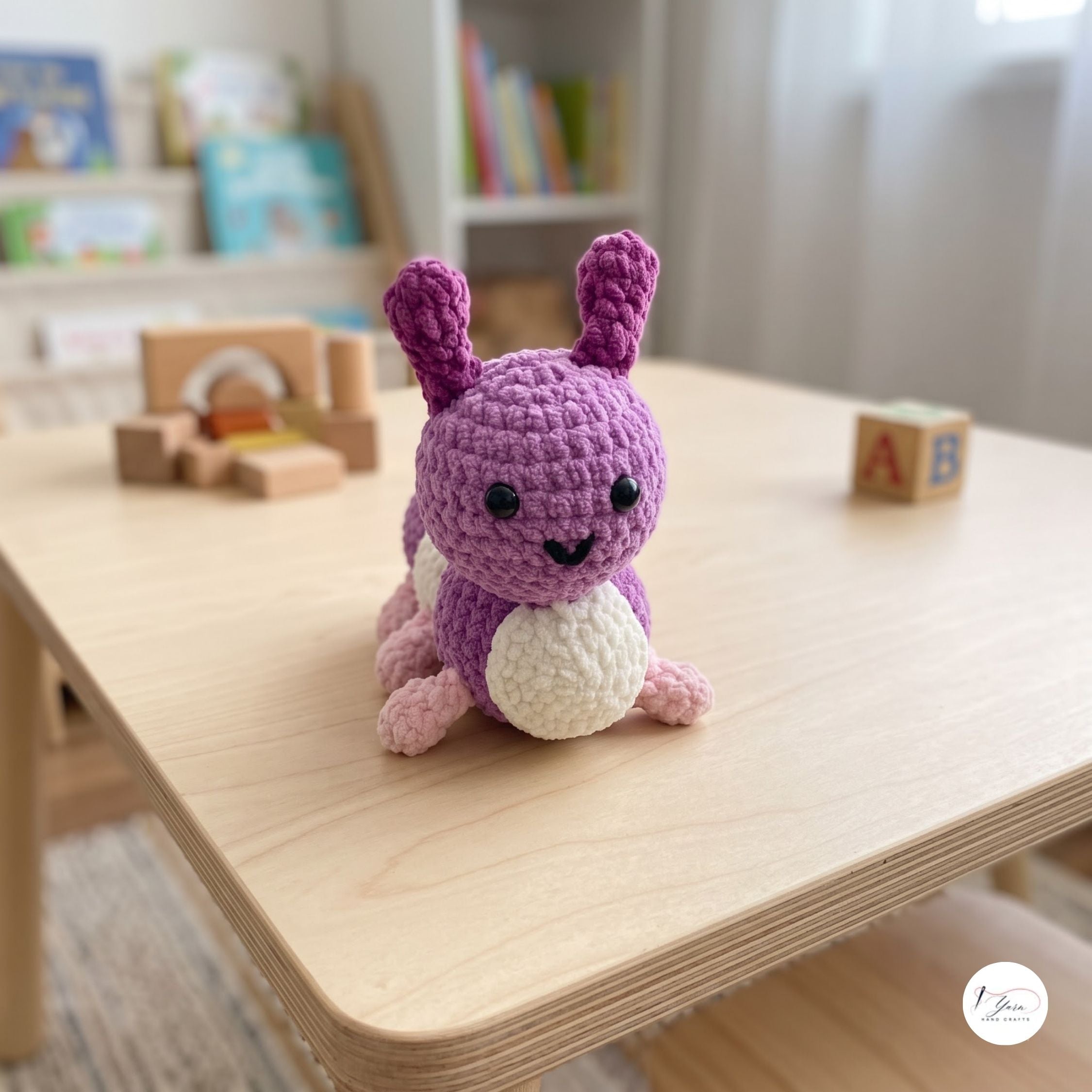 A purple and cream crocheted bunny toy sits on a light wood table in a softly blurred children's room setting.