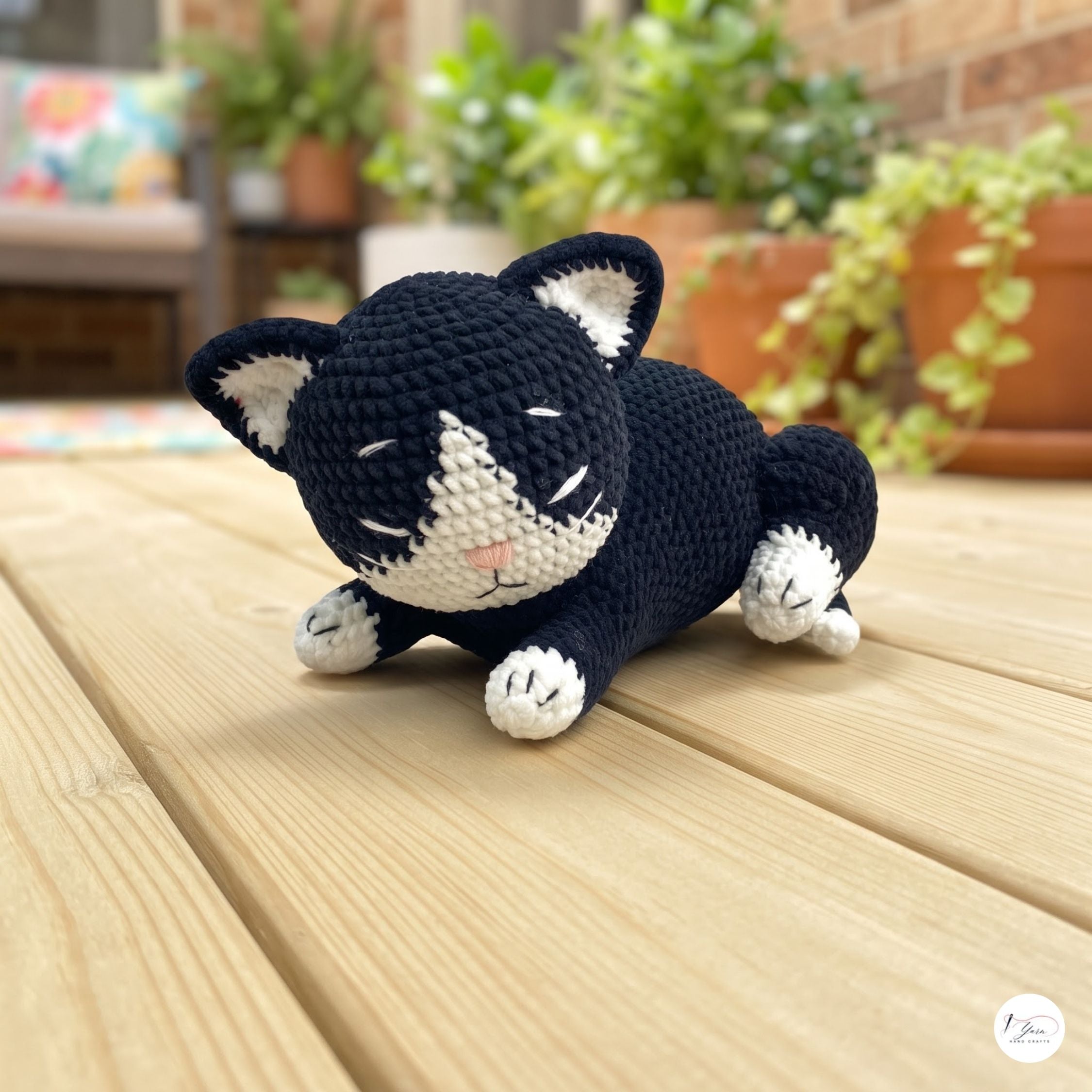 A crocheted sleeping cat toy in black and white, resting on a light wooden deck with a blurred garden background.