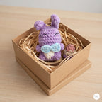 A crocheted purple bunny plush toy with a light blue bow and cream belly sits inside a brown kraft gift box filled with shredded paper on a light wood surface.