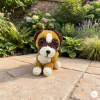 A handmade crocheted dog toy sits on a stone patio in a lush garden setting. The toy is brown and cream with black eyes and nose, and a small white circular logo is visible in the bottom right corner.