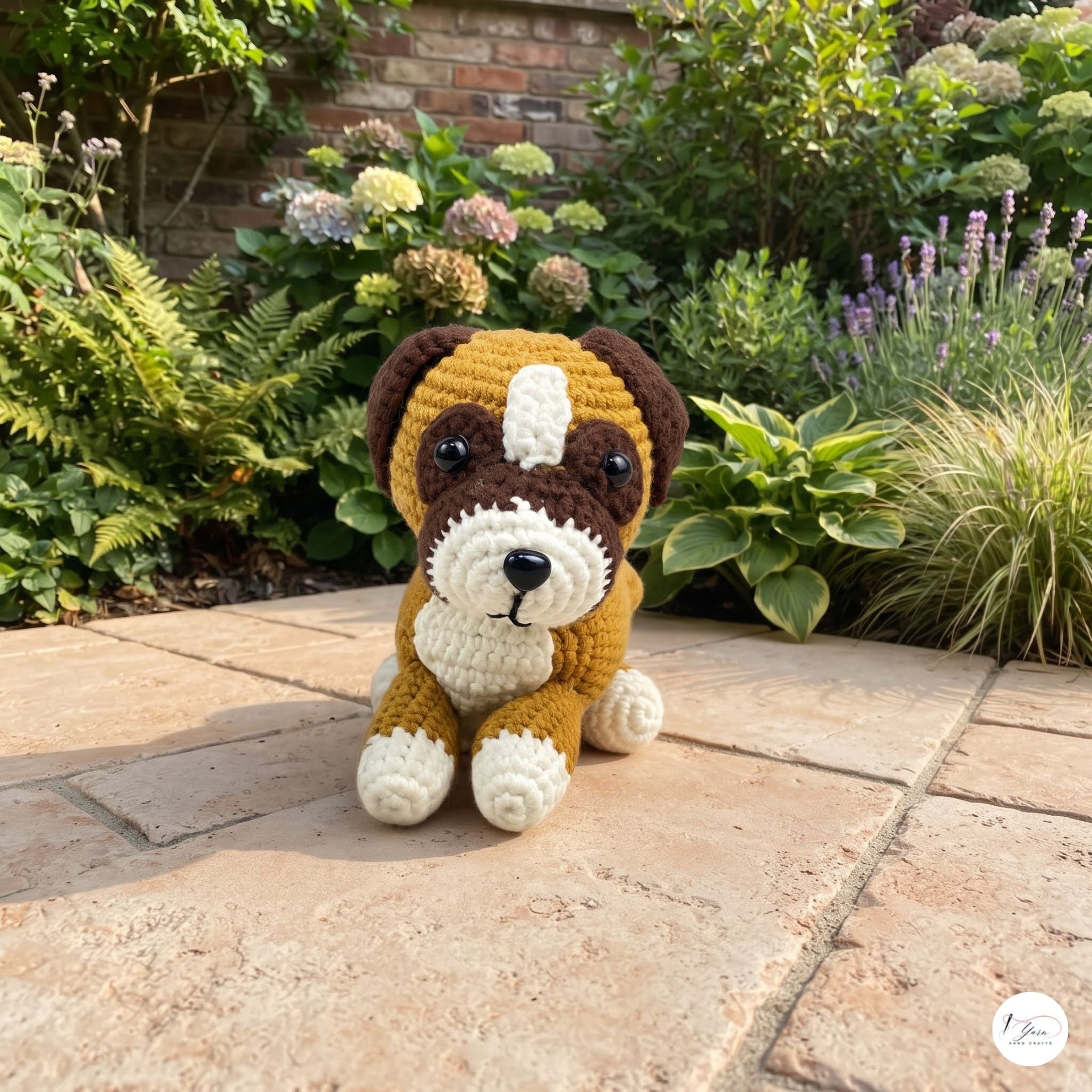 A handmade crocheted dog toy sits on a stone patio in a lush garden setting. The toy is brown and cream with black eyes and nose, and a small white circular logo is visible in the bottom right corner.