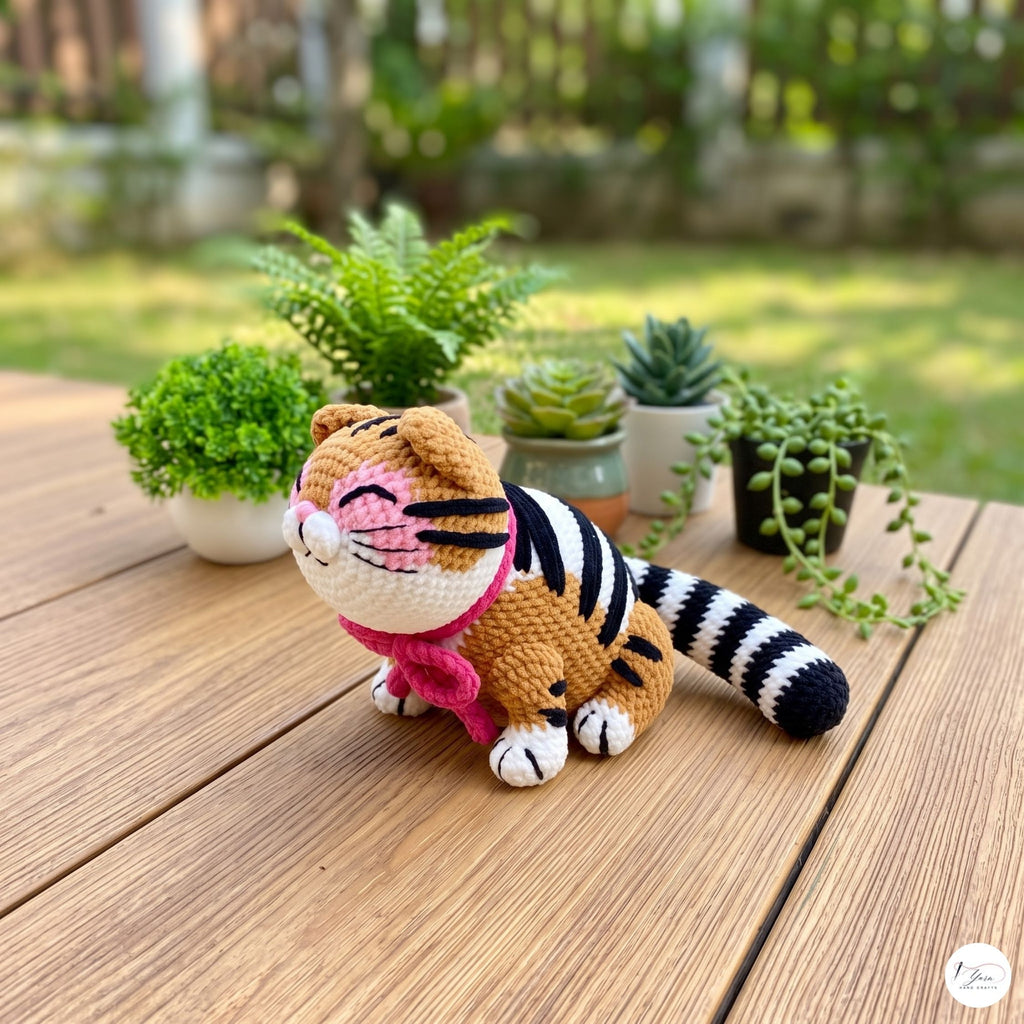 A crocheted tiger doll sits on a wooden table outdoors, with small potted plants and a blurred garden background. The doll has orange and black stripes, white paws and face details, and a pink collar.
