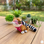 A crocheted tiger doll sits on a wooden table outdoors, with small potted plants and a blurred garden background. The doll has orange and black stripes, white paws and face details, and a pink collar.