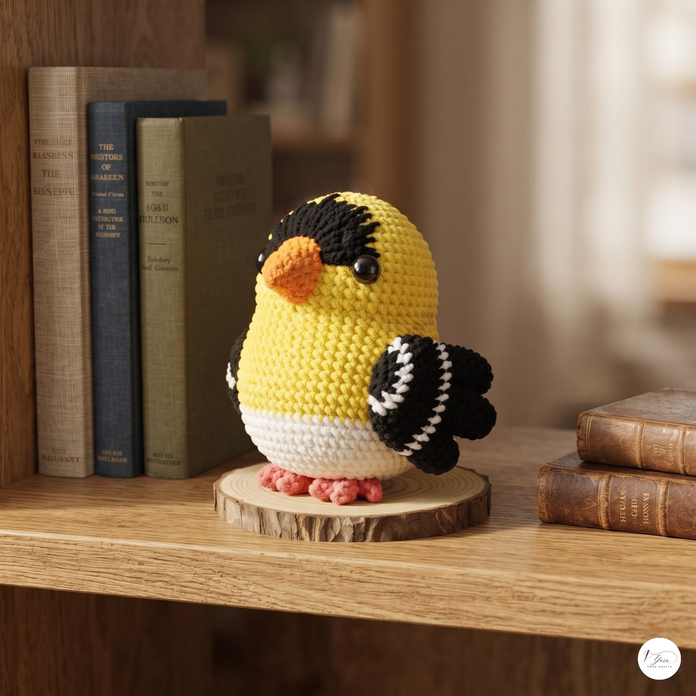 A crocheted goldfinch bird ornament displayed on a wooden slice stand on a wooden shelf, with books in the background.