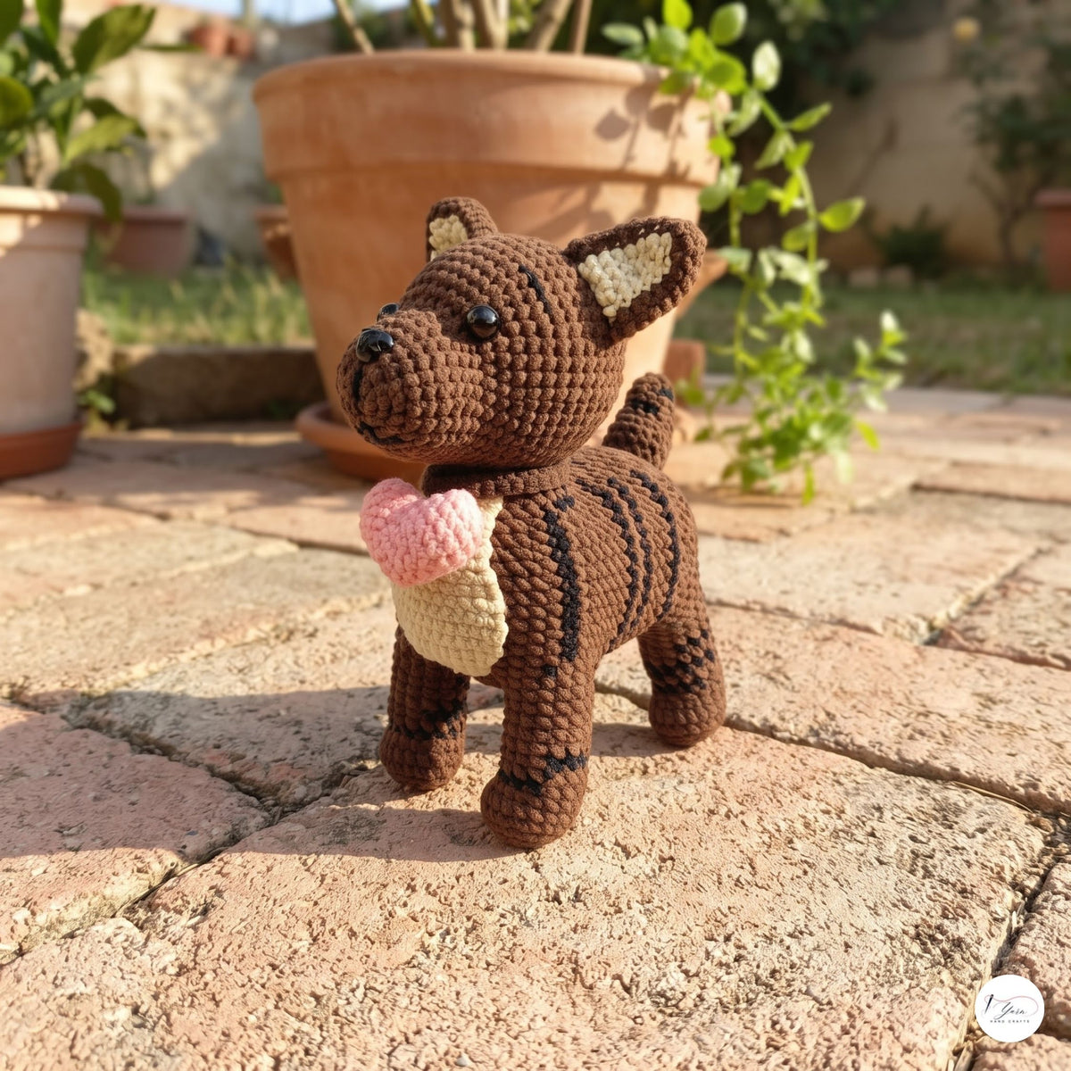 A crocheted plush dog toy with brown and black stripes, holding a pink and beige ice cream cone, standing on a stone patio in front of potted plants.