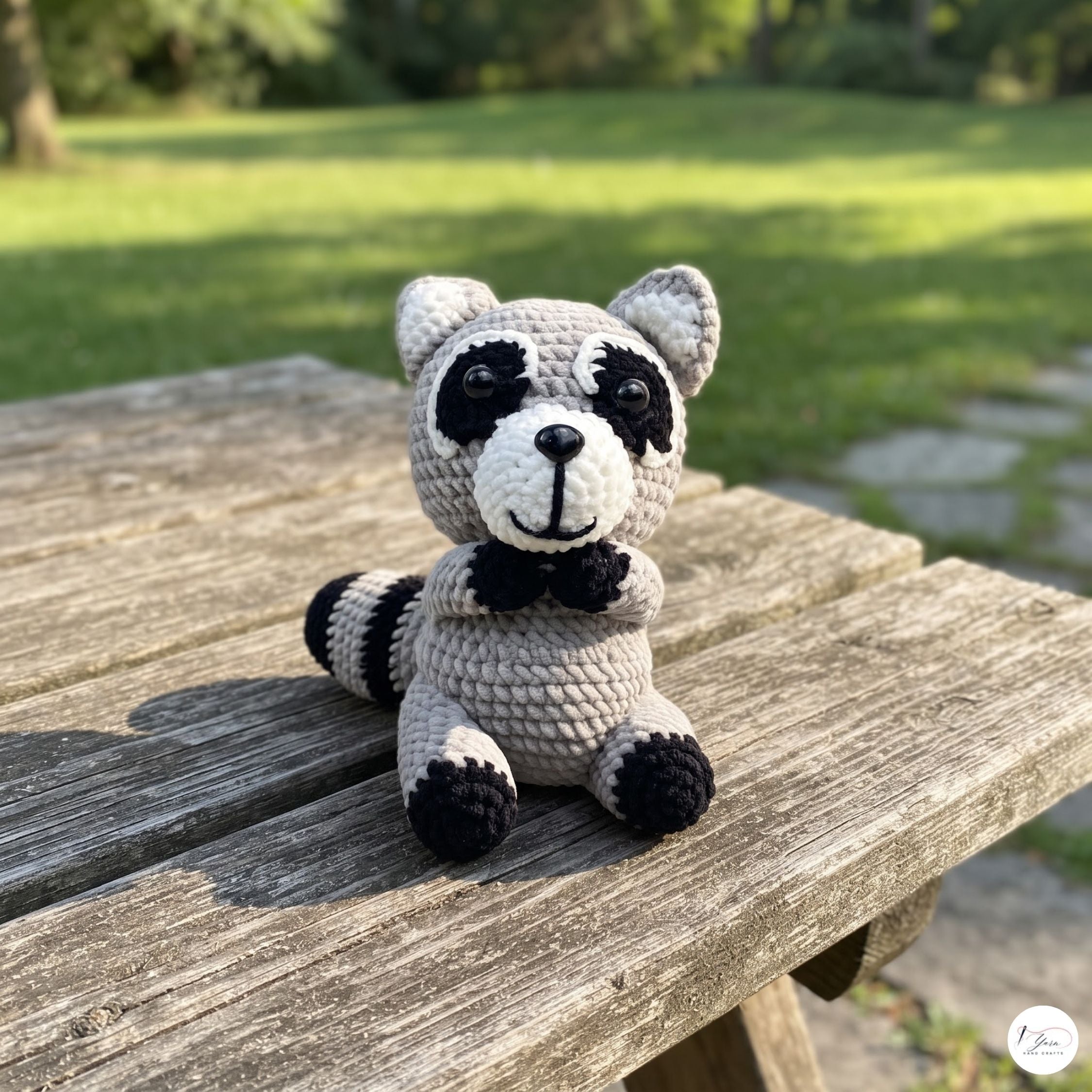 A crocheted raccoon toy sits on a weathered wooden picnic table outdoors, with a blurred green lawn and trees in the background.