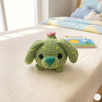 A green crocheted plush dog toy with yellow stitched details, a teal nose, and a small pink flower on its head, sitting on a white bed next to a children's book.