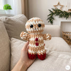 A hand holds a crocheted gingerbread doll with beige and white striped yarn, red buttons, and red feet, set against a cozy living room background with a beige sofa and holiday decorations.