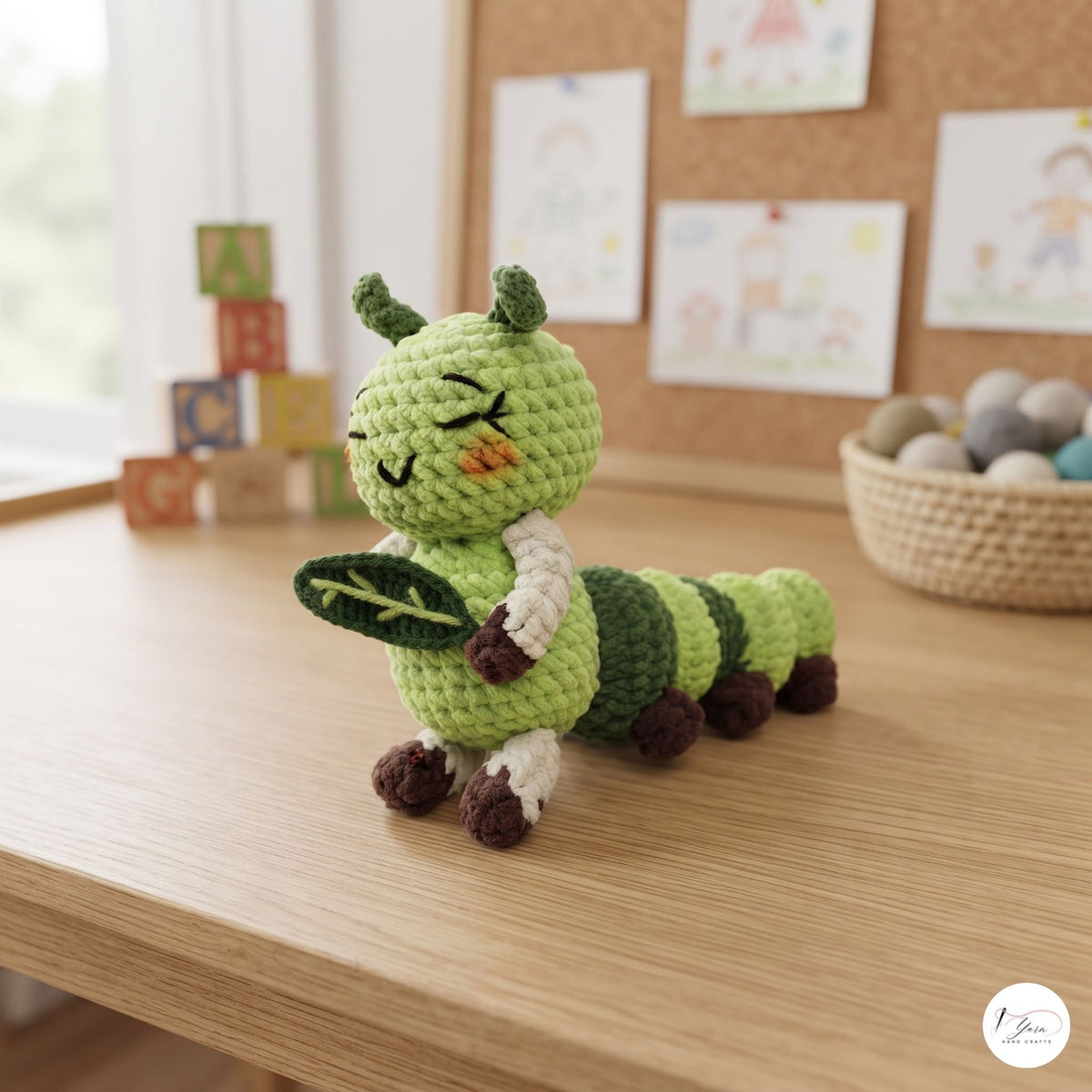 A crocheted green caterpillar toy holding a leaf, sitting on a light wooden table in a softly lit room with toys and framed pictures in the background.