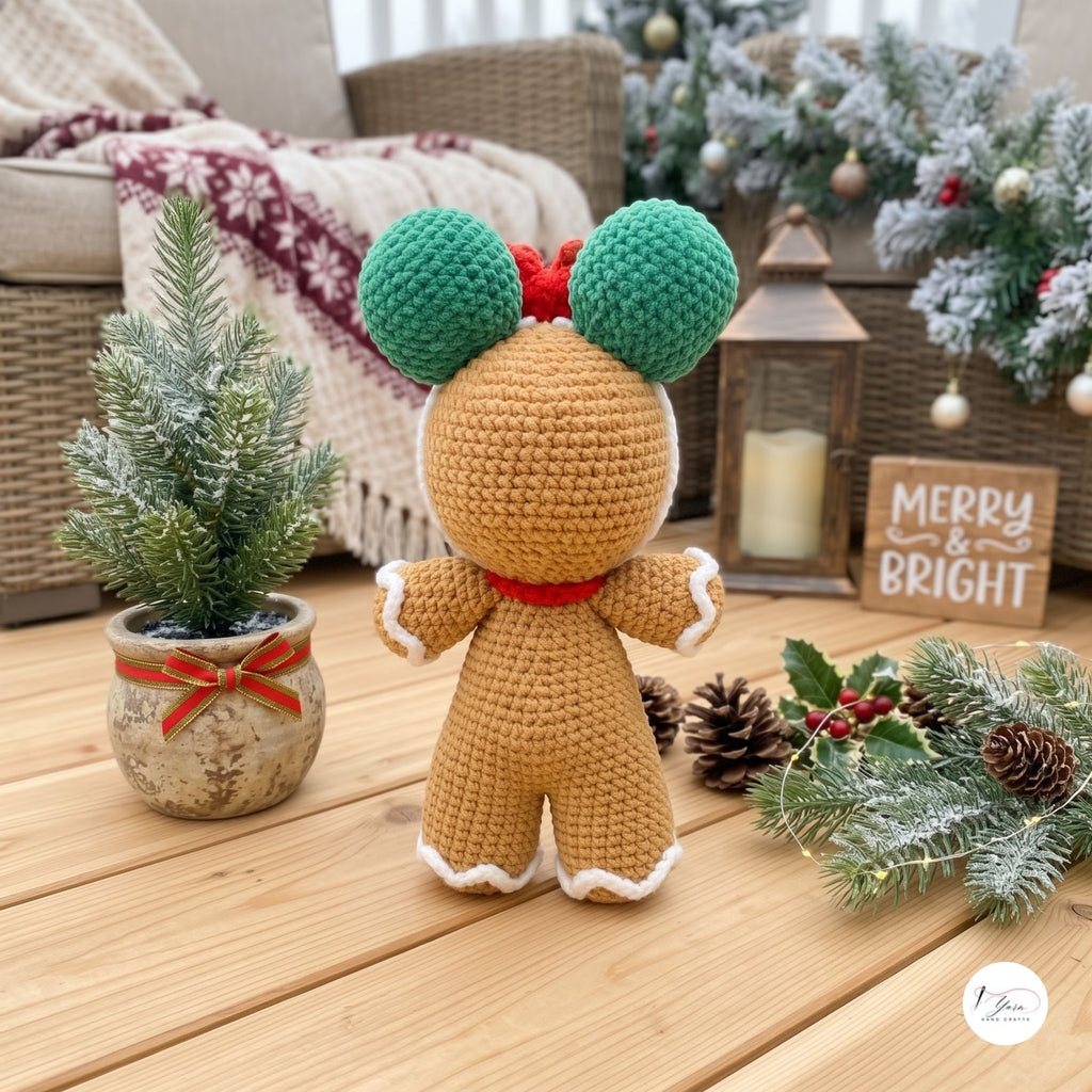 A crocheted gingerbread doll shown from the back, standing on a wooden floor in a cozy holiday setting with a small potted tree, wreath, lantern, and a 'Merry & Bright' sign.