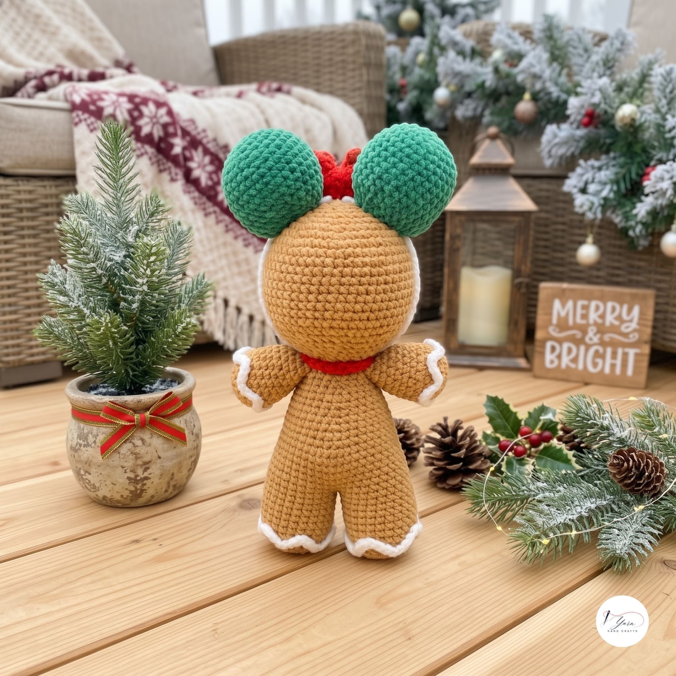 A crocheted gingerbread doll shown from the back, standing on a wooden floor in a cozy holiday setting with a small potted tree, wreath, lantern, and a 'Merry & Bright' sign.