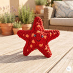 A red crocheted starfish toy with orange spots and a smiling face, standing on a light wooden table in an outdoor patio setting with potted plants and a sofa blurred in the background.