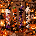 Four crocheted Halloween hanging ornaments displayed on a rustic branch in a cozy, warmly lit fall setting. The ornaments include a white ghost with orange pumpkin accents, an orange jack-o'-lantern, a black cat with purple witch hat and yellow moon details, and a white mummy with black spider accents.