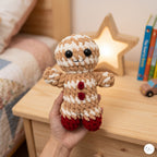 A hand holds a crocheted gingerbread person doll in a cozy bedroom setting with a wooden nightstand, a glowing star lamp, and books in the background.