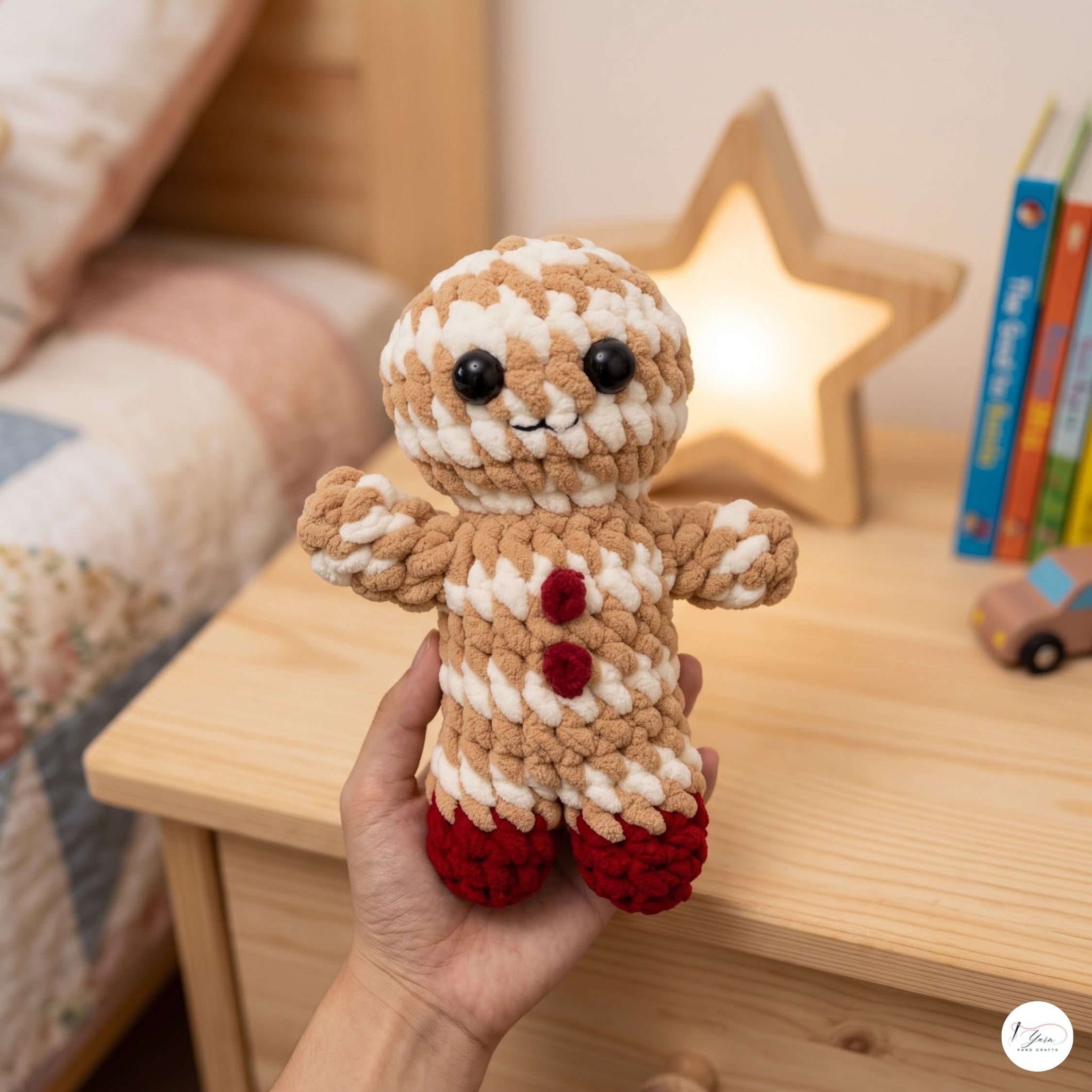 A hand holds a crocheted gingerbread person doll in a cozy bedroom setting with a wooden nightstand, a glowing star lamp, and books in the background.