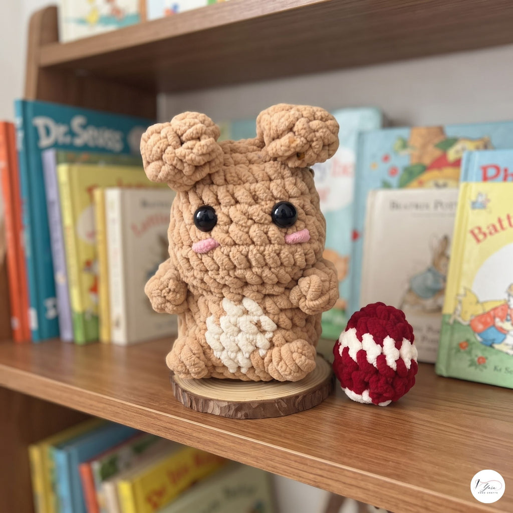 A crocheted amigurumi bunny figure in tan and cream sits on a wooden bookshelf next to a small red-and-white crocheted egg. Books are visible in the background on the shelf.