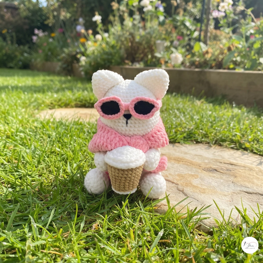 A cute handmade white crochet cat plush sitting on green grass in an outdoor garden, wearing a pink sweater, dark pink-framed sunglasses over its eyes, and holding a crocheted takeaway coffee cup.