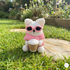 A cute handmade white crochet cat plush sitting on green grass in an outdoor garden, wearing a pink sweater, dark pink-framed sunglasses over its eyes, and holding a crocheted takeaway coffee cup.