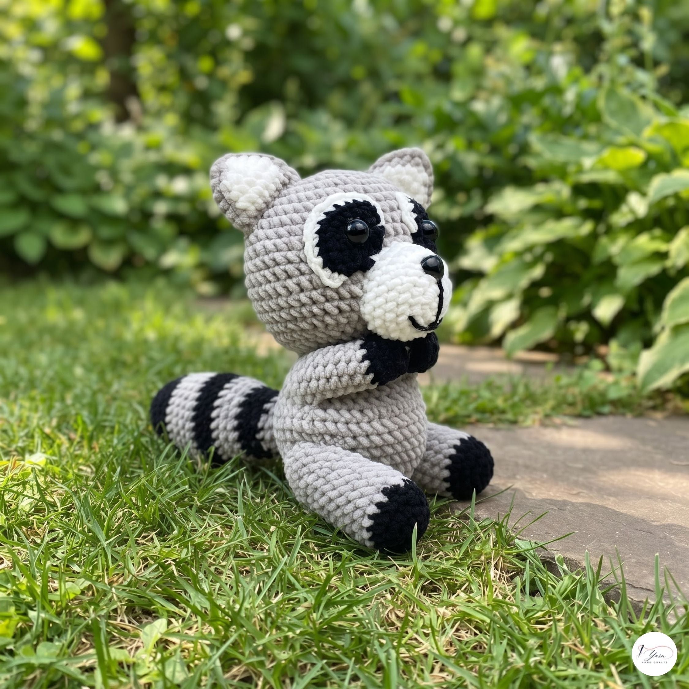 A crocheted raccoon toy sits on green grass outdoors with a blurred garden background. The toy is made of light gray and white yarn with black accents on the eyes, ears, paws, and tail stripes.