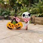 Two handmade crocheted Halloween-themed toys on a stone patio with a garden background. One toy features two orange jack-o'-lanterns with brown stems and pink loops, and the other features a white skull on a brown stem with pink and mint green loops.