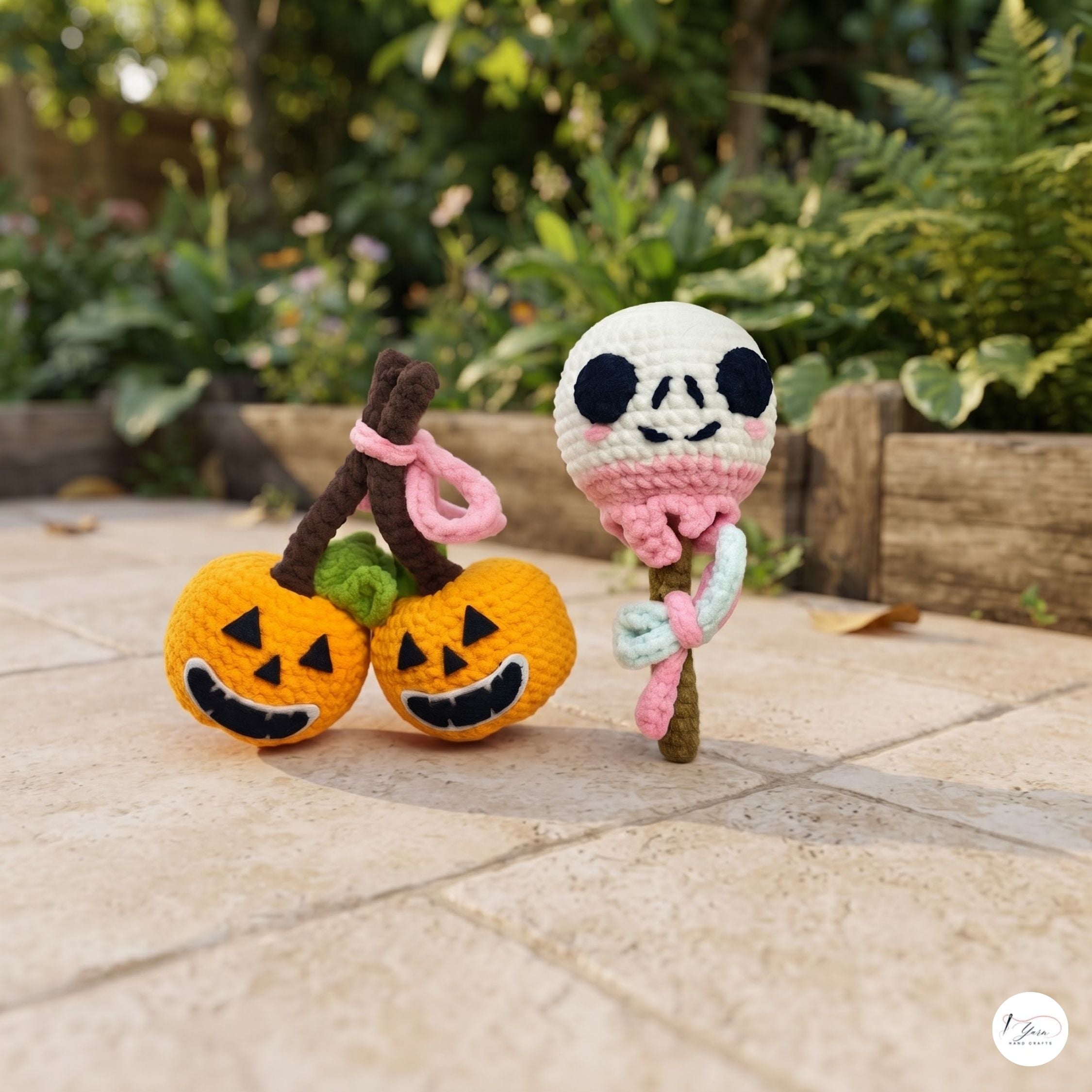 Two handmade crocheted Halloween-themed toys on a stone patio with a garden background. One toy features two orange jack-o'-lanterns with brown stems and pink loops, and the other features a white skull on a brown stem with pink and mint green loops.