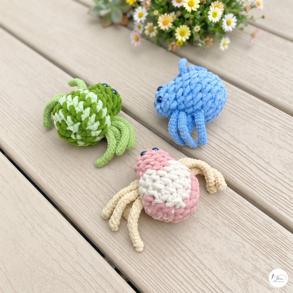 Three crocheted crab toys in green-and-white, blue, and pink-and-cream are arranged on a light wood plank surface with small daisies in the background.