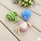 Three crocheted crab toys in green-and-white, blue, and pink-and-cream are arranged on a light wood plank surface with small daisies in the background.