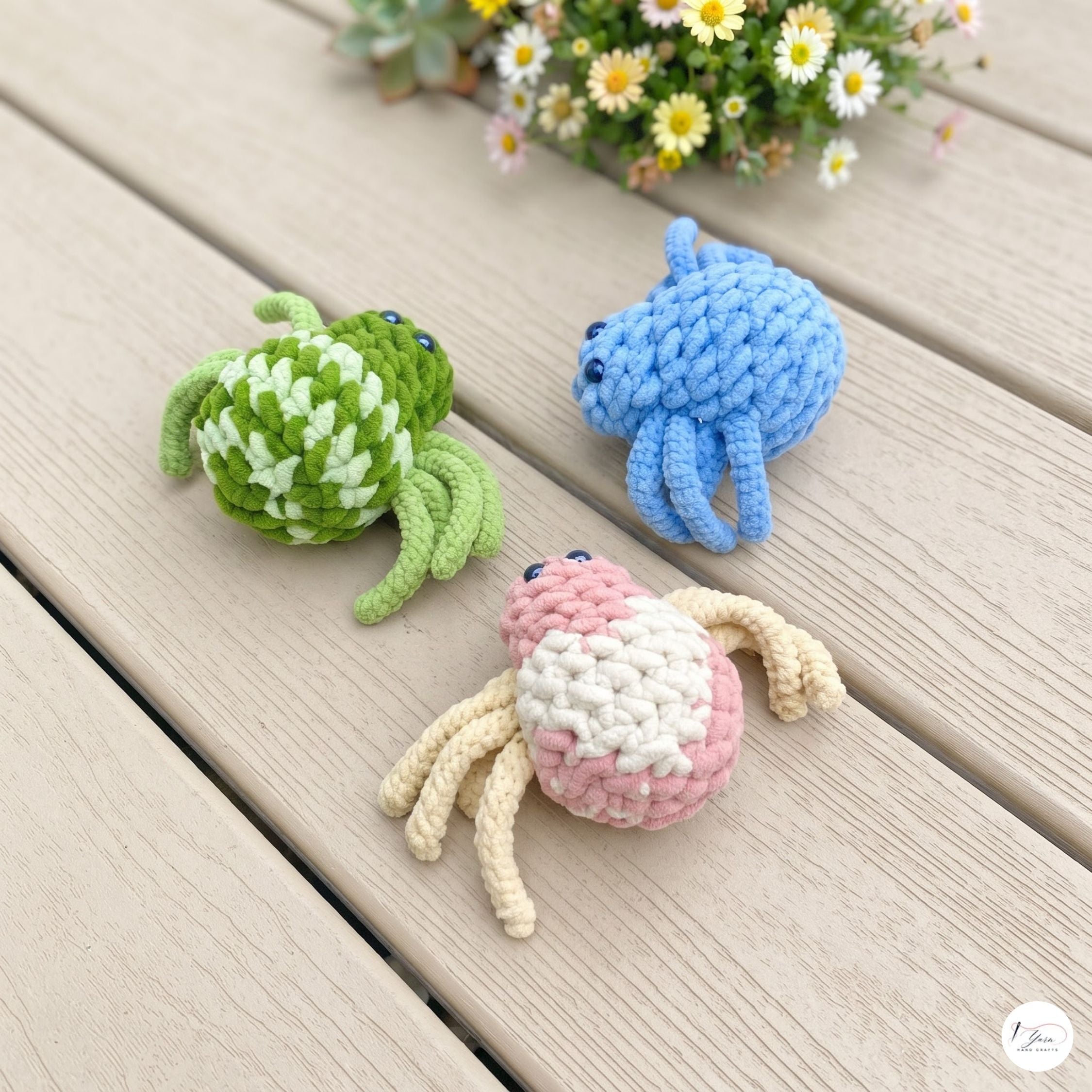 Three crocheted crab toys in green-and-white, blue, and pink-and-cream are arranged on a light wood plank surface with small daisies in the background.
