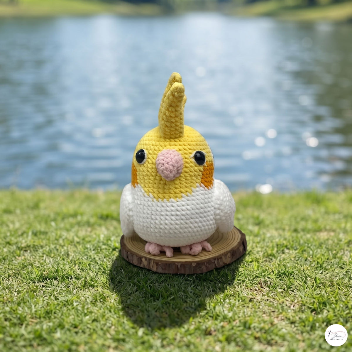 A crocheted cockatiel toy standing on a small round wooden slice, placed on green grass with a blurred lake and hills in the background.