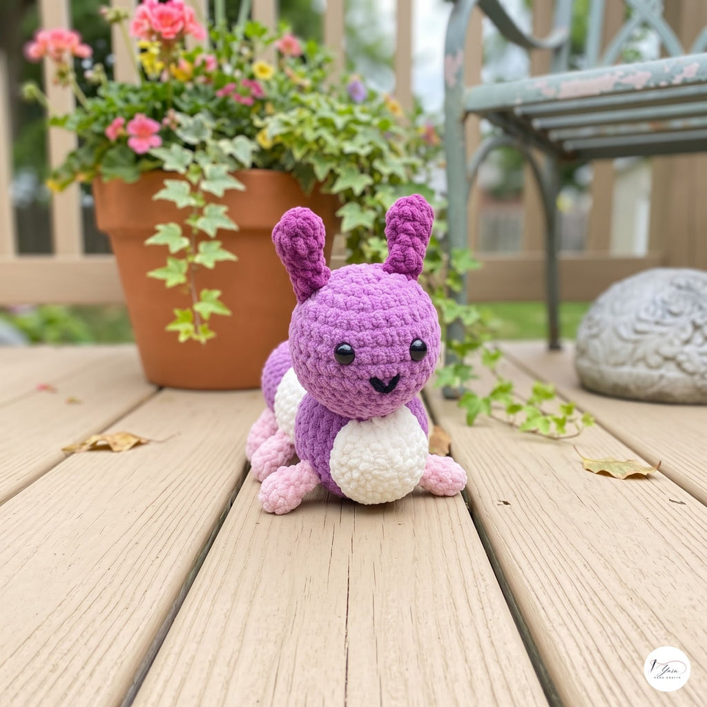 A small crocheted bunny toy sits on a wooden deck outdoors, with a potted flowering plant and a garden chair blurred in the background.