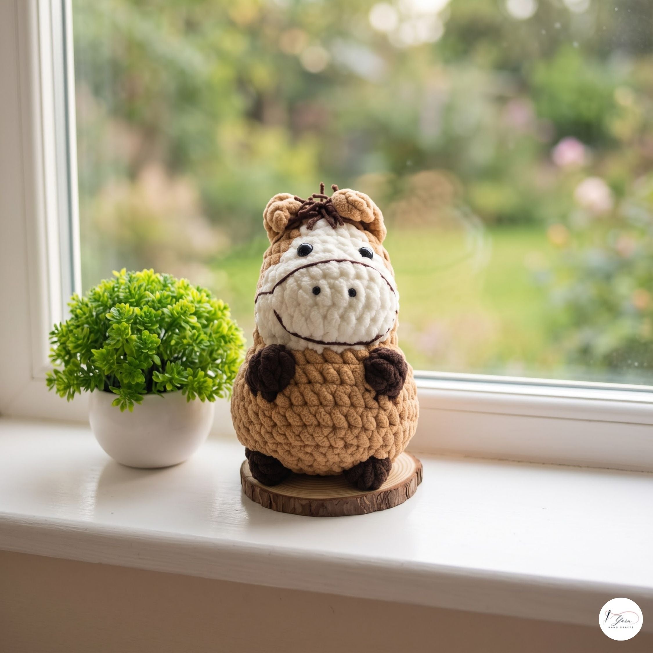 A crocheted cow amigurumi toy sits on a wooden coaster on a white windowsill, next to a small potted green plant. The background shows a softly blurred green garden view through the window.