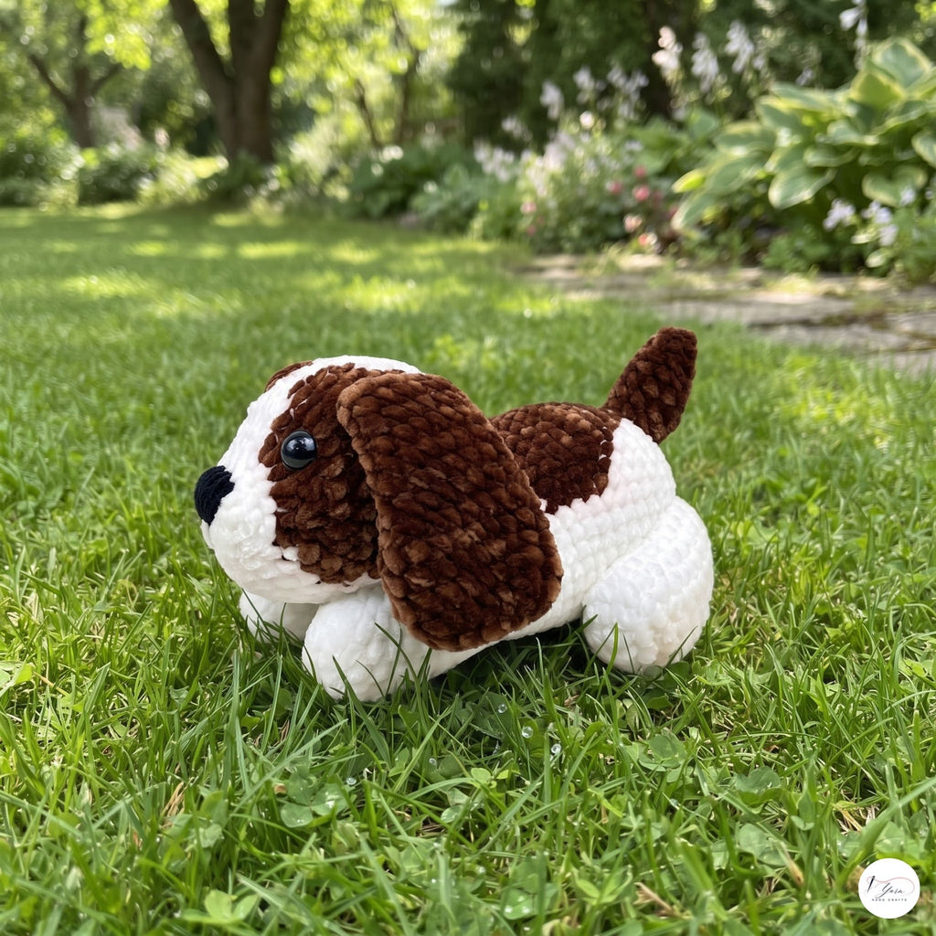A crocheted plush puppy toy with brown and white yarn, lying on a green grass lawn with a blurred garden background.