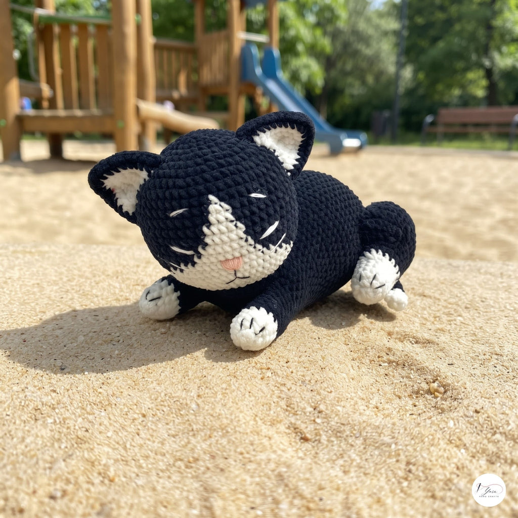 A crocheted black-and-white cat toy lying on sand in an outdoor playground setting, with a wooden playset and trees blurred in the background.