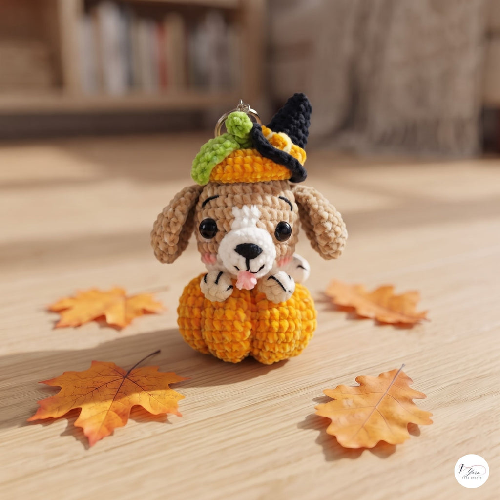 A crocheted dog-shaped keychain or charm dressed in a pumpkin costume, sitting on a wooden surface with scattered autumn leaves. The dog has tan and white yarn, a yellow pumpkin body, and a black and green witch hat.