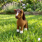 A crocheted toy horse standing on green grass in a garden, with flowers and plants blurred in the background.