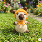 A crocheted amigurumi bear doll wearing a yellow pumpkin hat and matching scarf, sitting on a green grassy lawn with a blurred garden background.