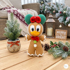 A crocheted gingerbread woman doll with green hat, red bow, yellow beak, and white trim stands on a wooden table in a cozy holiday setting with a small potted tree, lantern, pinecones, and a 'Merry & Bright' sign.