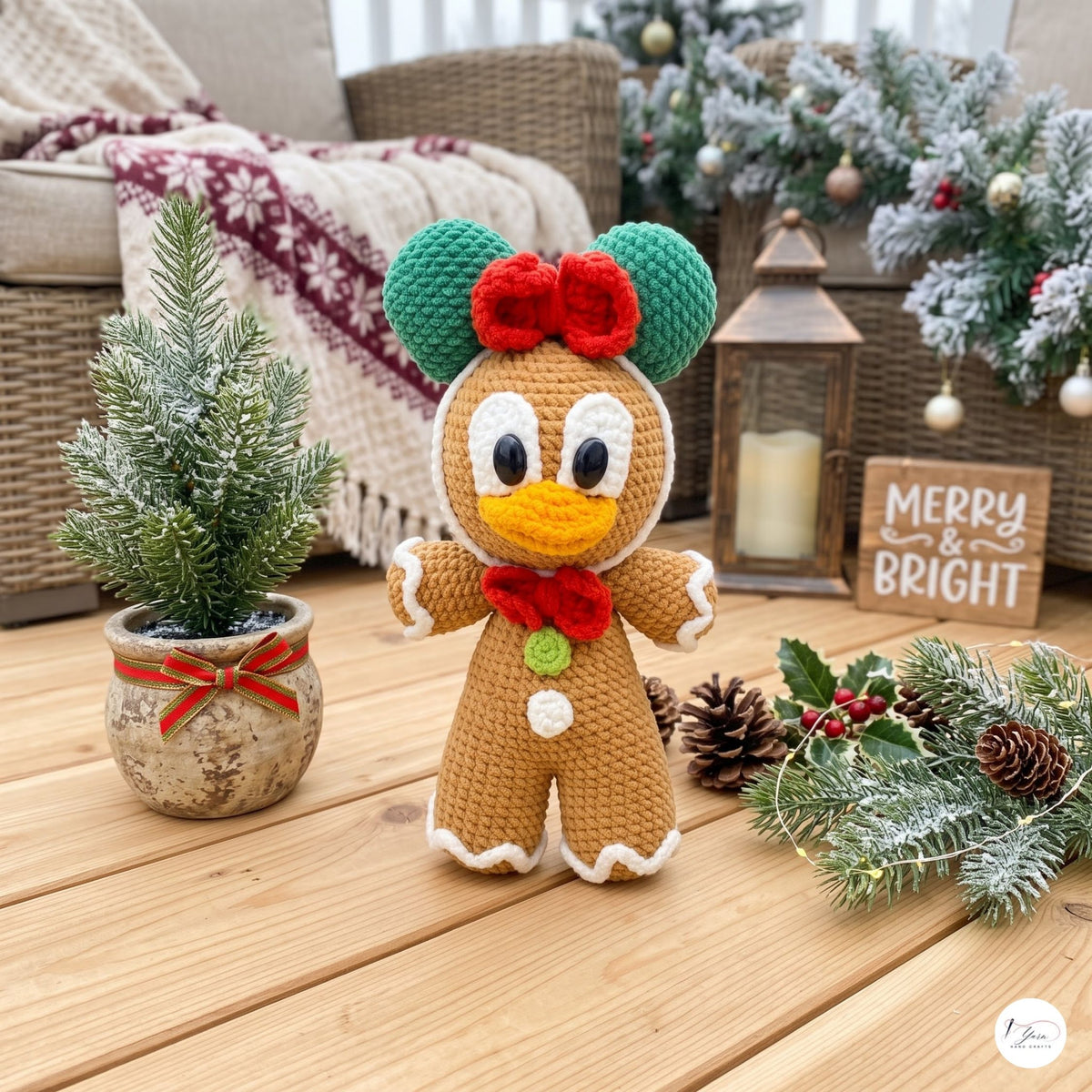 A crocheted gingerbread woman doll with green hat, red bow, yellow beak, and white trim stands on a wooden table in a cozy holiday setting with a small potted tree, lantern, pinecones, and a 'Merry & Bright' sign.