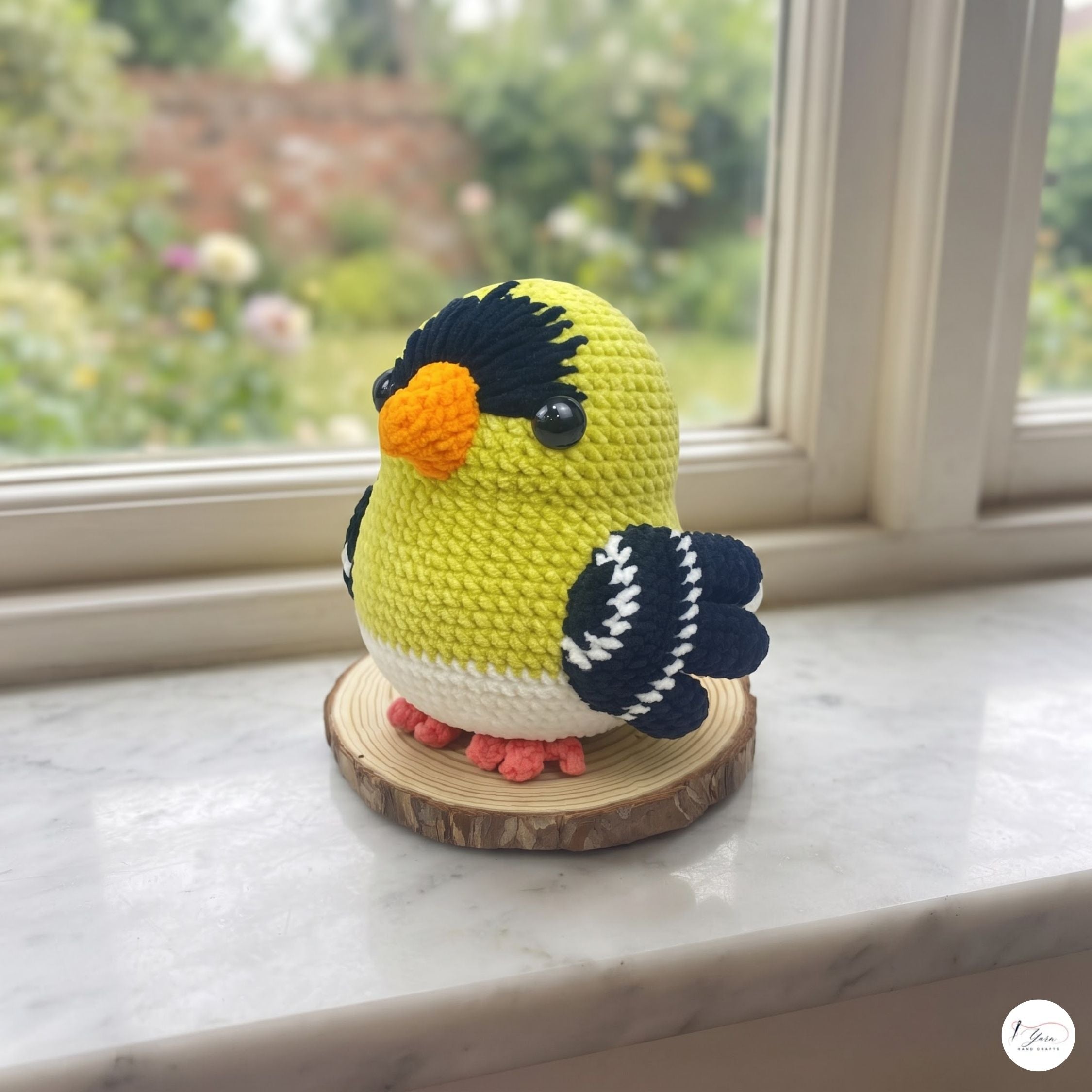 A crocheted bird figurine with a yellow body, black and white wings, and an orange beak, standing on a round wooden slice base on a marble windowsill with a garden view in the background.