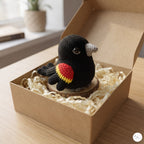 A crocheted black bird figurine with a gray beak and a red, yellow, and black wing detail, displayed inside a brown kraft gift box filled with shredded paper on a wooden table.