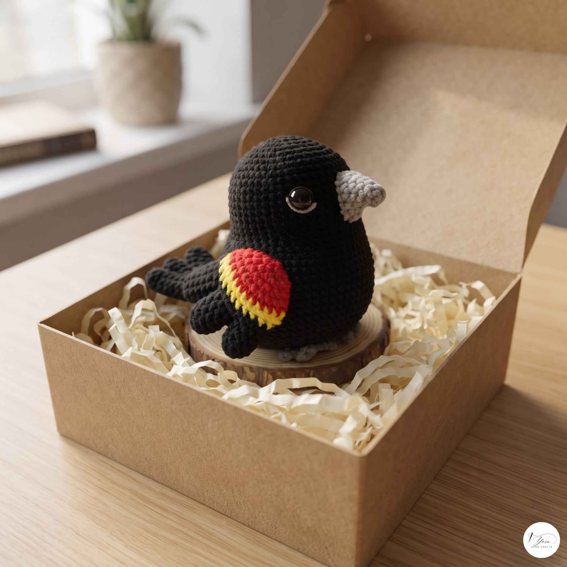 A crocheted black bird figurine with a gray beak and a red, yellow, and black wing detail, displayed inside a brown kraft gift box filled with shredded paper on a wooden table.