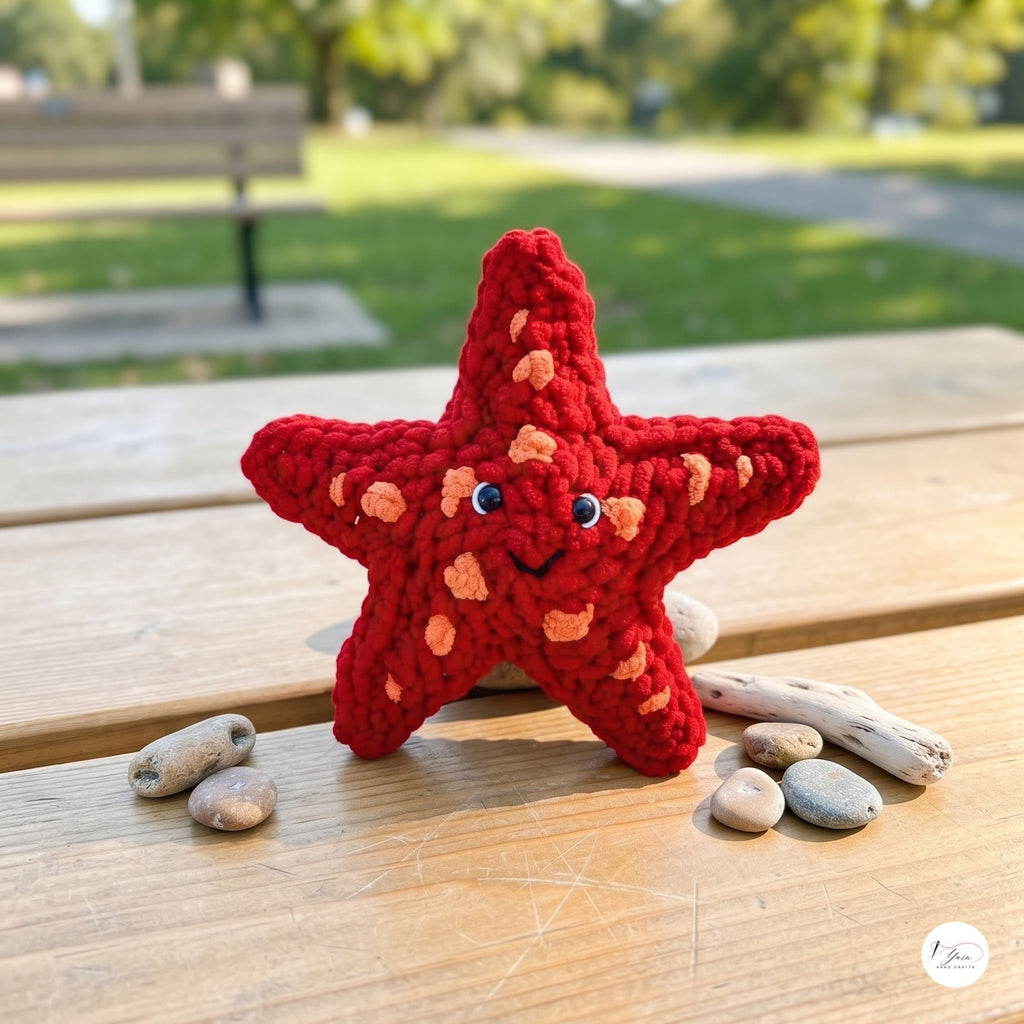 A red crocheted starfish toy with a smiling face and small blue eyes sits on a wooden table outdoors, surrounded by small stones and driftwood, with a blurred park background.