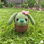 A plush handmade crochet cactus puppy toy with long floppy ears, yellow embroidered spines, a pink flower, and a turquoise nose, sitting inside a brown crochet pot base on a green lawn outdoor